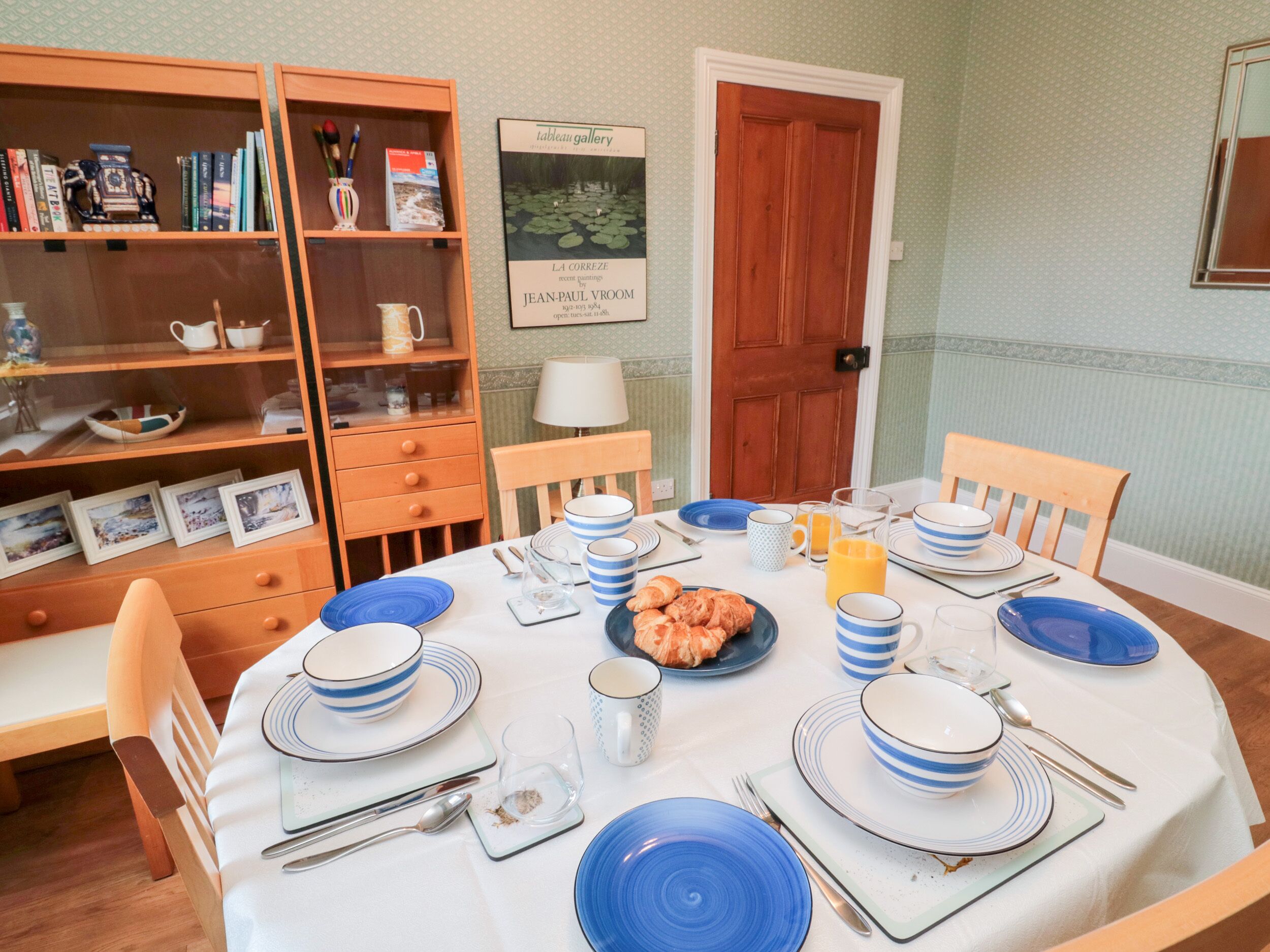 A dining area with a round table set for four, featuring blue and white plates, cups, and cutlery. There is a bookshelf to the left with books and decorative items. A framed picture hangs on the wall above the table.