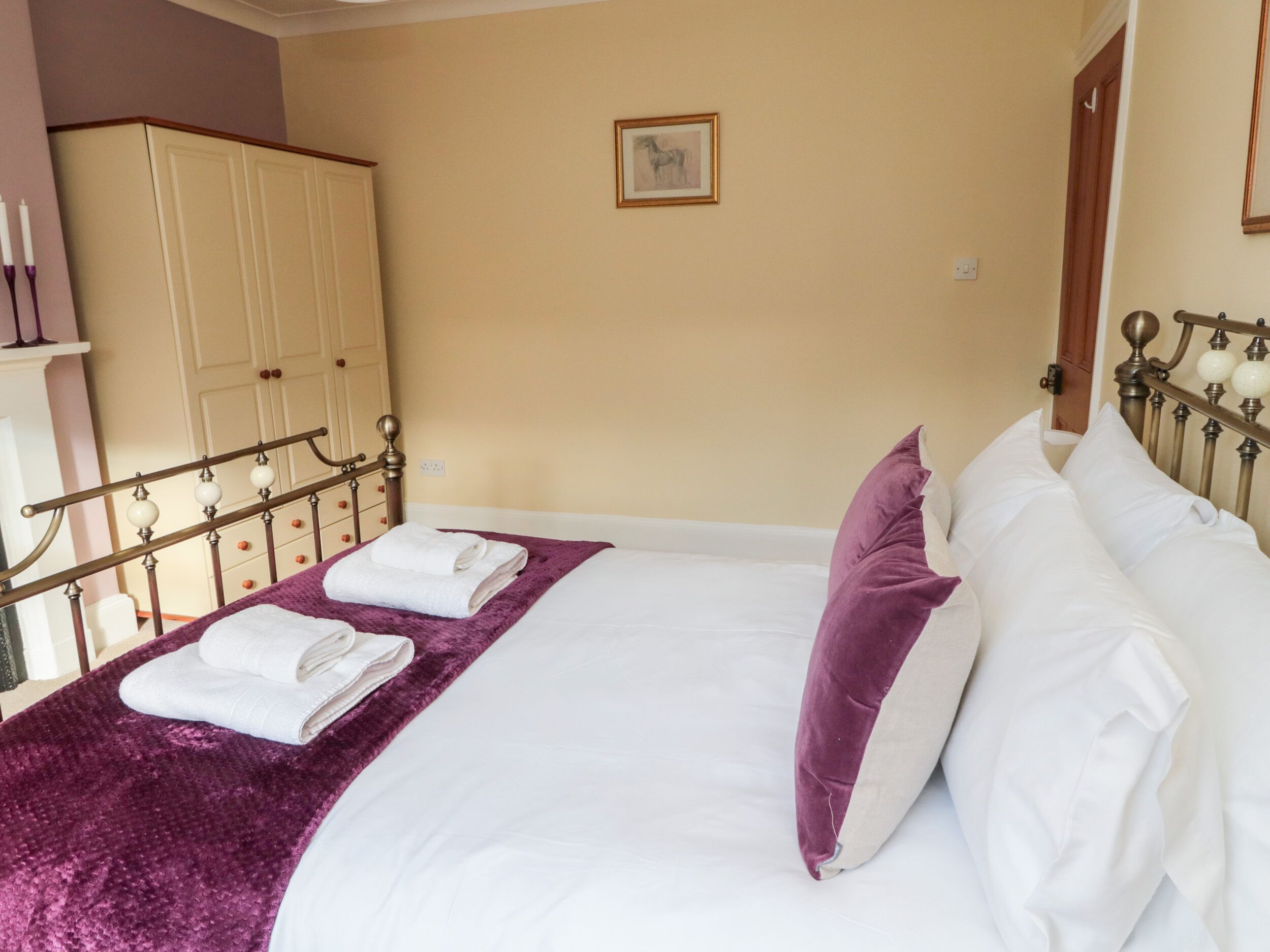 A neatly made bed with white linens and a purple throw blanket, a wooden wardrobe, and a framed picture on the wall.