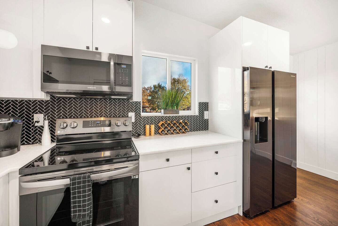 A modern kitchen with stainless steel appliances, white cabinetry, a herringbone backsplash, and a window with a view of trees.