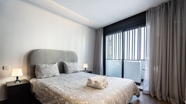 A modern hotel room with a large bed, two bedside tables with lamps, a view of the city through large windows, and a balcony. - Kifkif One (Bejaad)