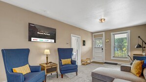 A living room with two blue armchairs, a grey sofa, a flat-screen TV displaying streaming services, and a window with a view of greenery. - Game Parlour! Downtown Workspace BBQ Fire Pit (Colorado Springs)