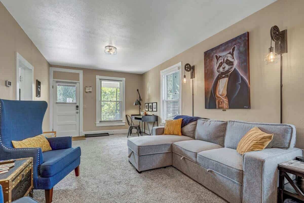 Ein Wohnzimmer mit blauem Sessel, grauem Ecksofa und Schreibtisch mit Stuhl. An der Wand hängt ein Bild von einem Waschbären.