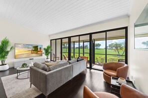 A modern living room with a grey sofa, a brown leather chair, and a large window offering a view of a green landscape. - New PGA Home on Fazio Golf Course w/ Amazing 180º Views by Guaranteed Rental™ (Palm Beach Gardens)