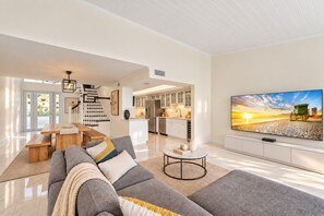 A spacious living room with a large flat-screen TV, a sofa, a dining area, and a kitchen in the background.