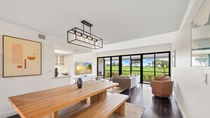 A modern living room with a wooden dining table, a sofa, and a chair. There is a large window with a view of a green landscape.