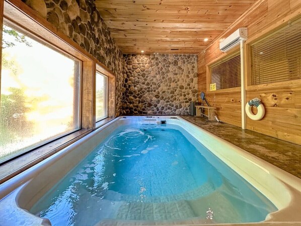 A hot tub in a room with stone walls and wooden ceiling, large windows, and a stone fireplace. - Gatlinburg Cabin w/ Indoor Heated Pool & Hot Tub (Gatlinburg)