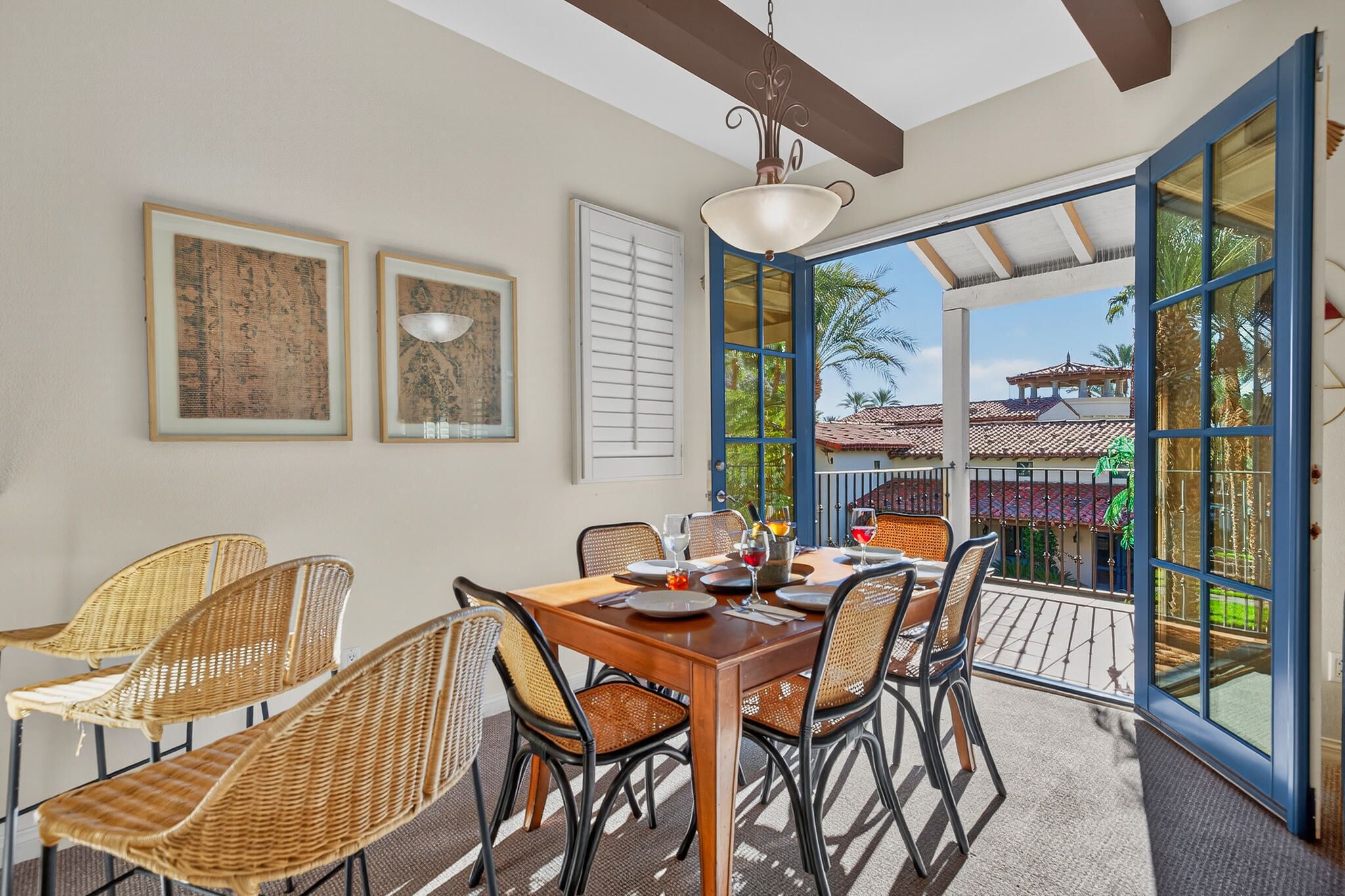 A dining area with a wooden table and wicker chairs, framed artwork on the wall, and a view of a traditional building through large glass doors.