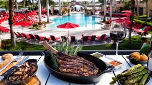 Una mesa de comedor con un filete a la parrilla, espárragos y una copa de vino tinto, con vistas a una zona de piscina con sombrillas y sillones de descanso rojos.