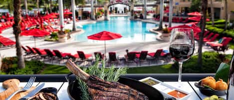 Ein Esstisch mit gegrilltem Steak, Spargel und einem Glas Rotwein, mit Blick auf einen Poolbereich mit roten Sonnenschirmen und Liegestühlen.