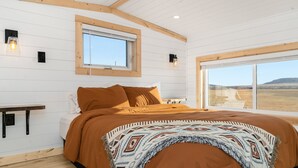 A bedroom with a large bed, wooden beams, a window, and a view of a landscape. - Modern Tiny Cabin, with Hot tub in Choteau MT (Choteau)