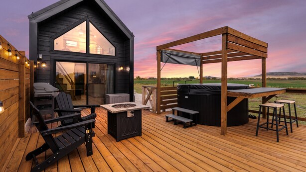 A wooden deck with a hot tub, seating area, and a view of the landscape. - Modern Tiny Cabin, with Hot tub in Choteau MT (Choteau)