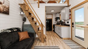 A compact living space with a wooden staircase, a kitchenette, a sofa with a brown cushion, and a view through a glass door. - Modern Tiny Cabin, with Hot tub in Choteau MT (Choteau)