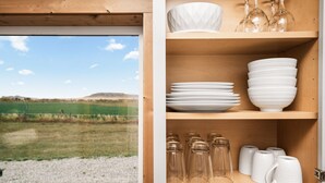 A wooden cabinet with shelves holding white dishes and glassware, a sign with a message, and a view of a grassy field through a window. - Modern Tiny Cabin, with Hot tub in Choteau MT (Choteau)