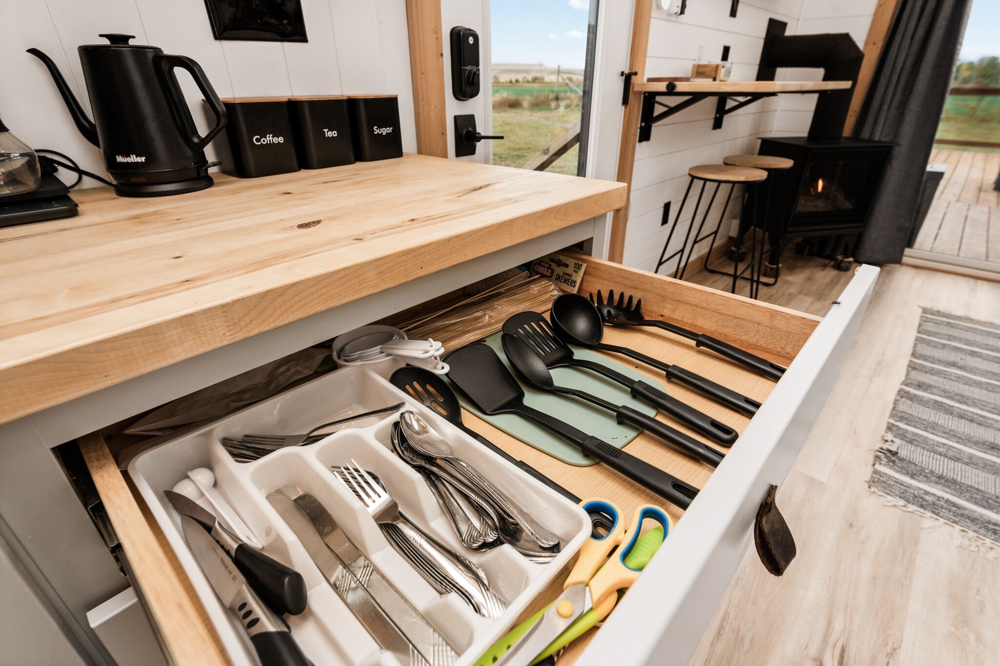 Una cucina con piano di lavoro in legno, una macchina da caffè, contenitori per zucchero e tè, e un cassetto con utensili.