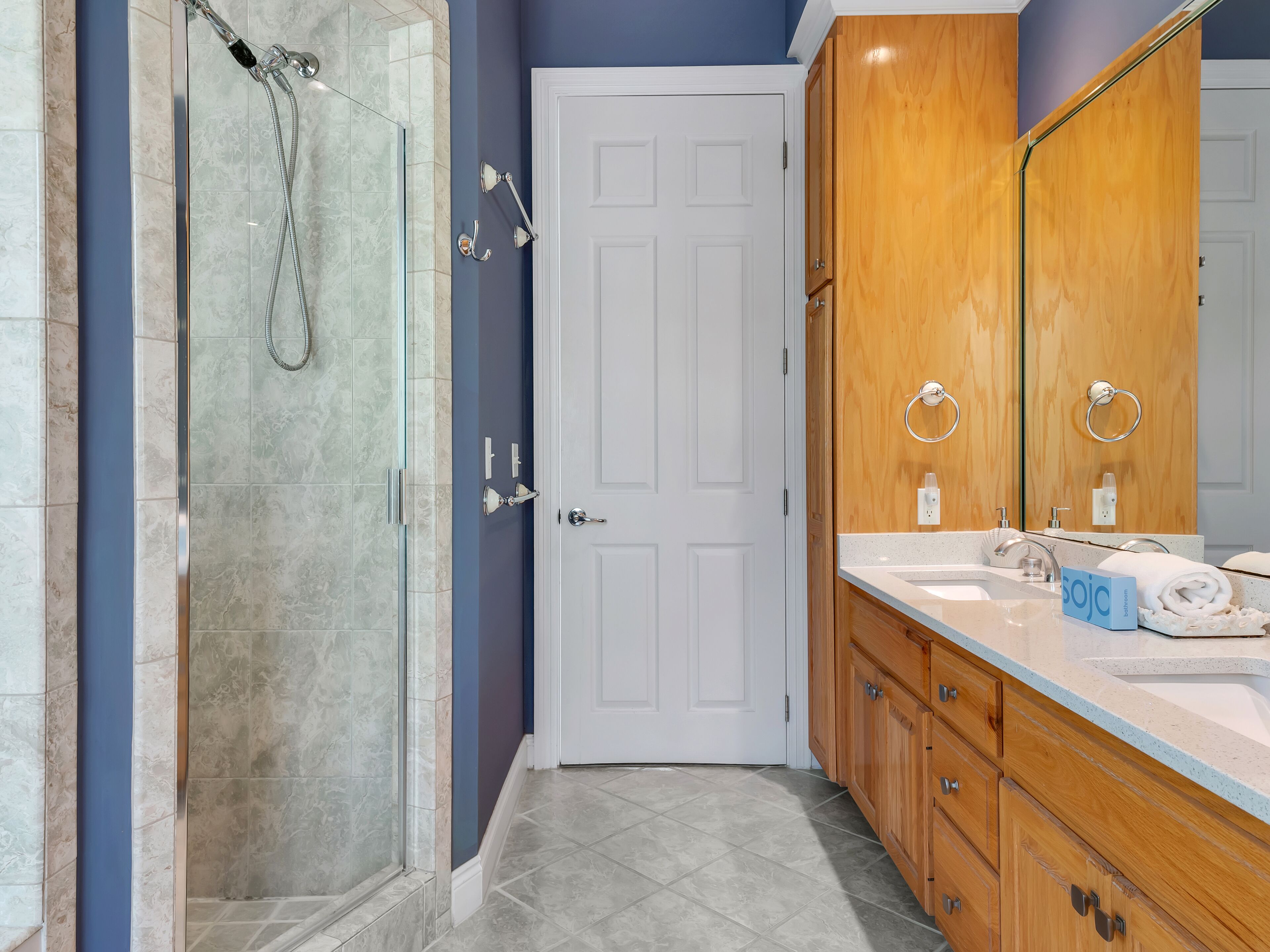 A bathroom with a glass shower enclosure, a white door, wooden cabinets, and a sink with a soap dispenser.