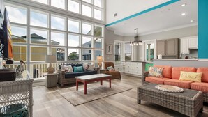 A spacious living room with large windows, a kitchen in the background, and a view of neighboring houses.