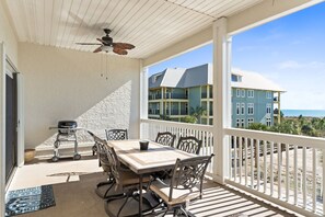 A covered outdoor patio with a table and chairs, a ceiling fan, and a view of buildings and the beach.