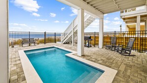 A rectangular pool with a white border, surrounded by a brick patio. There is a covered area with a white ceiling and a staircase leading to an upper level.
