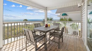 A covered outdoor area with a wooden table and chairs, a view of the beach, and a building with a white facade and green roof.