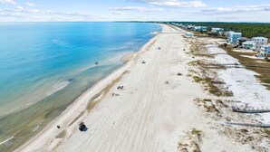 An aerial view of a beach with a clear sky, a sandy shore, and a row of buildings along the coastline.