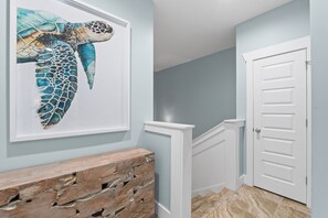 Un pasillo con un banco de madera, una puerta blanca y una obra de arte enmarcada de una tortuga marina.
