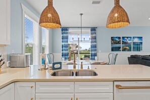Una cucina con una grande isola, due lampade a sospensione in vimini, un lavello incassato e vista sull'esterno attraverso una finestra con tende blu.