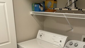 Un cuarto de lavado con lavadora y secadora de carga frontal, un estante con productos de limpieza y una puerta.