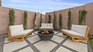 An outdoor seating area with a central fire pit, surrounded by two wooden chairs and a bench, set against a brick wall with tall, narrow plants. - This Must Be The Place ! (Phoenix)