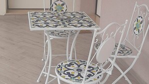 Una stanza con una porta bianca, un tavolino con tovaglia fantasia e due sedie in metallo bianco con schienali decorati.