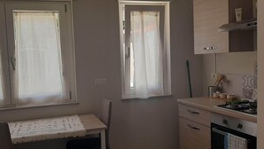 Una piccola cucina con un fornello a gas, un lavandino e una finestra con una tenda bianca. C'è un piccolo tavolo con una tovaglia bianca e una sedia.