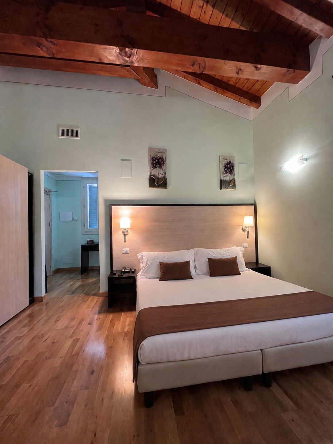 Een slaapkamer met een houten plafond, een groot bed met wit en bruin beddengoed, twee wandlampen en een klein nachtkastje met een lamp.
