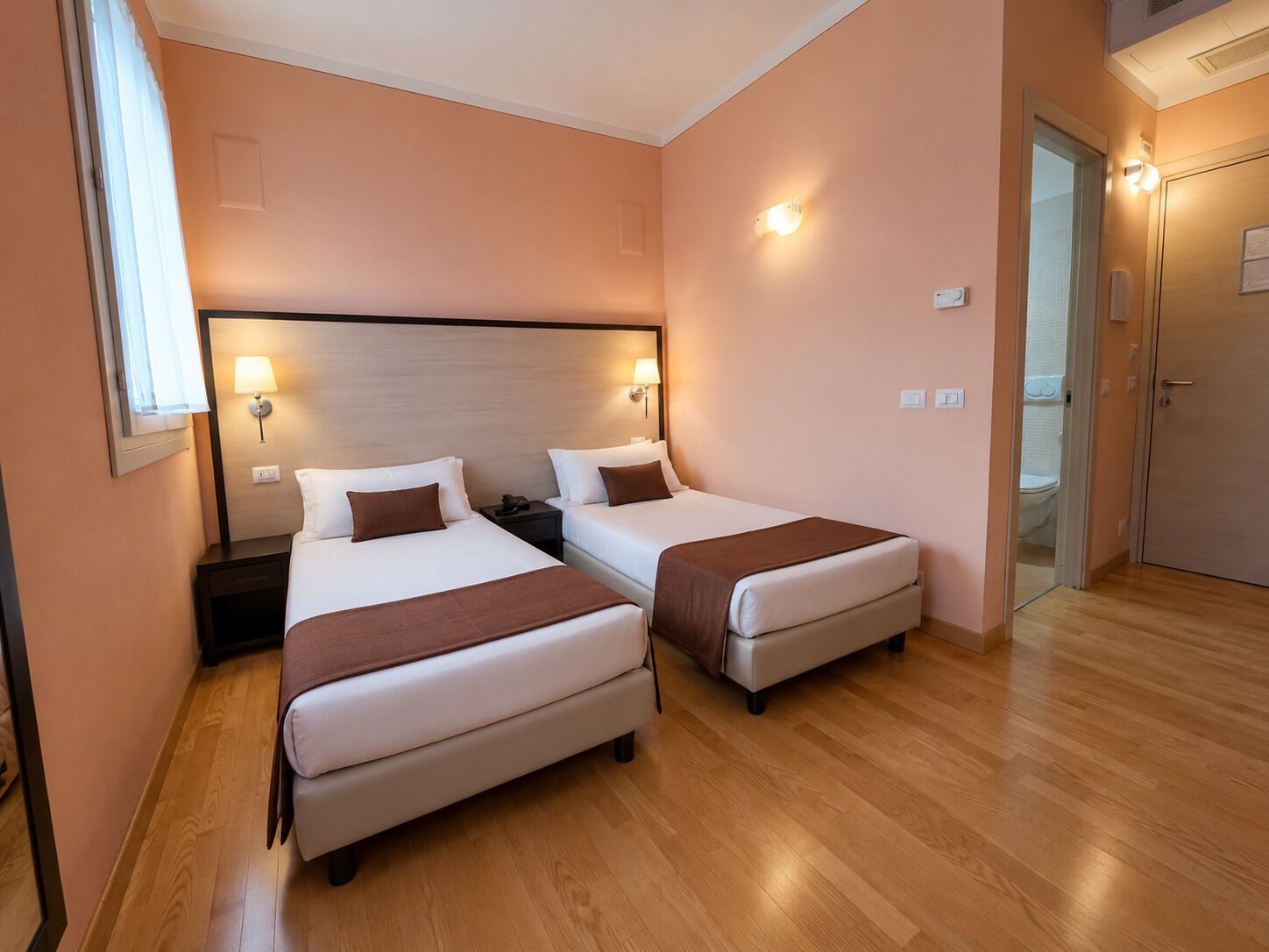 A hotel room with two beds, wooden flooring, a nightstand with a lamp, and a door leading to a bathroom.