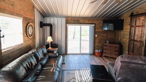 Interior acogedor de una cabaña con sofá de cuero, techo de madera, televisor y puerta corrediza de vidrio que da a un espacio exterior.