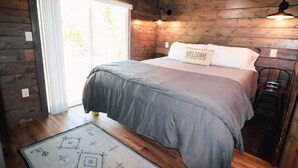 Una habitación tipo cabaña de madera con una cama, un letrero de bienvenida y una pequeña mesa con un radio.
