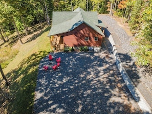 A wooden cabin with a sloped roof, surrounded by a gravel driveway and a wooden fence. - Stunning Mtn Views, Hot Tub+Fire Pit, WiFi, Grill! (Bakersville)