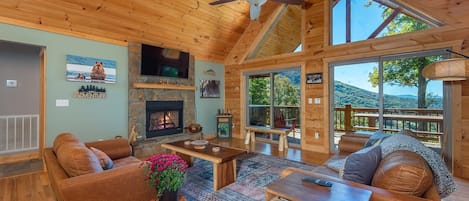 A spacious living room with a fireplace, wooden furniture, and large windows offering a view of the outdoors.