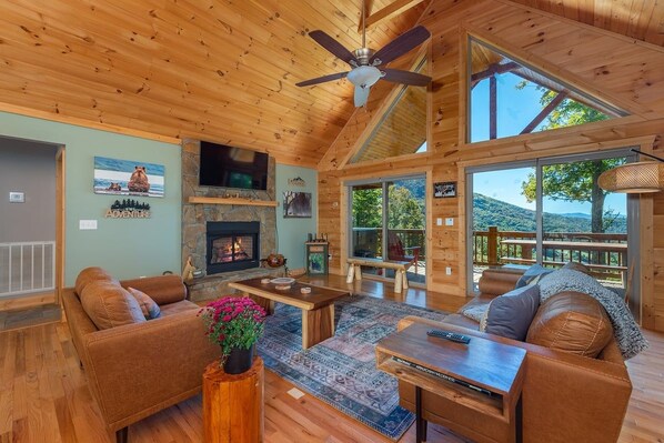 A spacious living room with a fireplace, wooden furniture, and large windows offering a view of the outdoors. - Stunning Mtn Views, Hot Tub+Fire Pit, WiFi, Grill! (Bakersville)