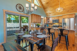 A spacious kitchen with wooden cabinets, a dining area with a table set for four, and a large window overlooking a stone wall. - Stunning Mtn Views, Hot Tub+Fire Pit, WiFi, Grill! (Bakersville)