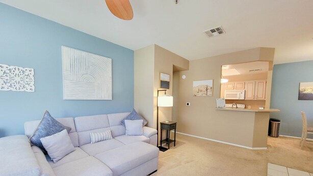 A living room with a light-colored sectional sofa, a small side table, and a kitchen area with a microwave and cabinets. - Private North Scottsdale Condo - Pool, Spa, Sauna & Outdoor Kitchen (Scottsdale)