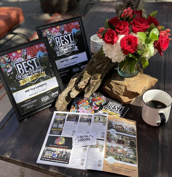 Des certificats encadrés récompensant le gagnant du prix « Meilleur établissement de River Valley 2023 », un bouquet de roses rouges, une tasse à café et des brochures sur l’établissement Hill Top Lodging.