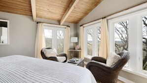 A bedroom with a large bed, two wicker chairs, a small table with a lamp, and large windows with curtains. - Mountain Forest View Lodge (Hackett)