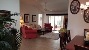 Un salon avec un canapé rouge, une table basse et une table à manger avec des chaises. On y trouve un ventilateur de plafond, une lampe et un luminaire fixé au mur.
