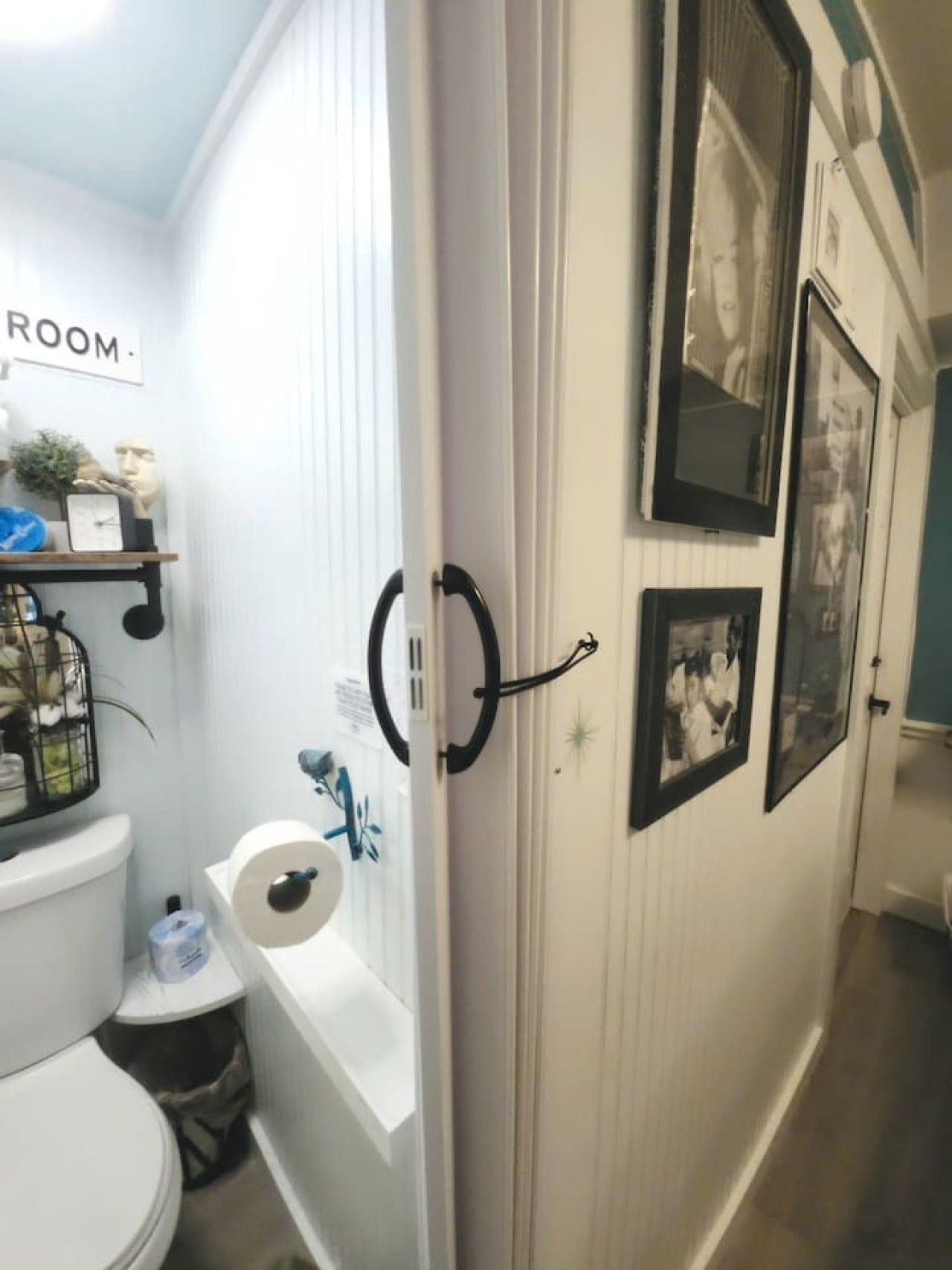 Een badkamer met een toilet, een rol toiletpapier, een kleine prullenbak en een plank met een klok en een plant.