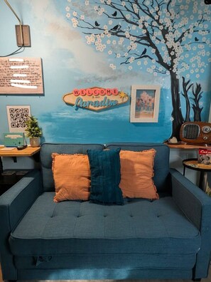 A blue sofa with two pillows, a wall with a 'Welcome to Paradise' sign, a QR code, and a vintage radio. - Island Parlor Inn Coastal Mid Century Studio (Savannah)