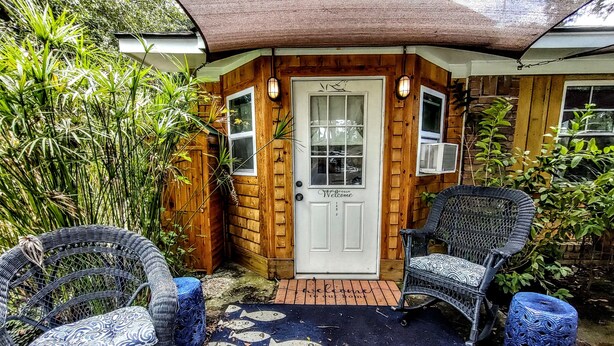 Une maison en bois avec une porte blanche, deux chaises en osier et un tapis orné de motifs de poissons.
