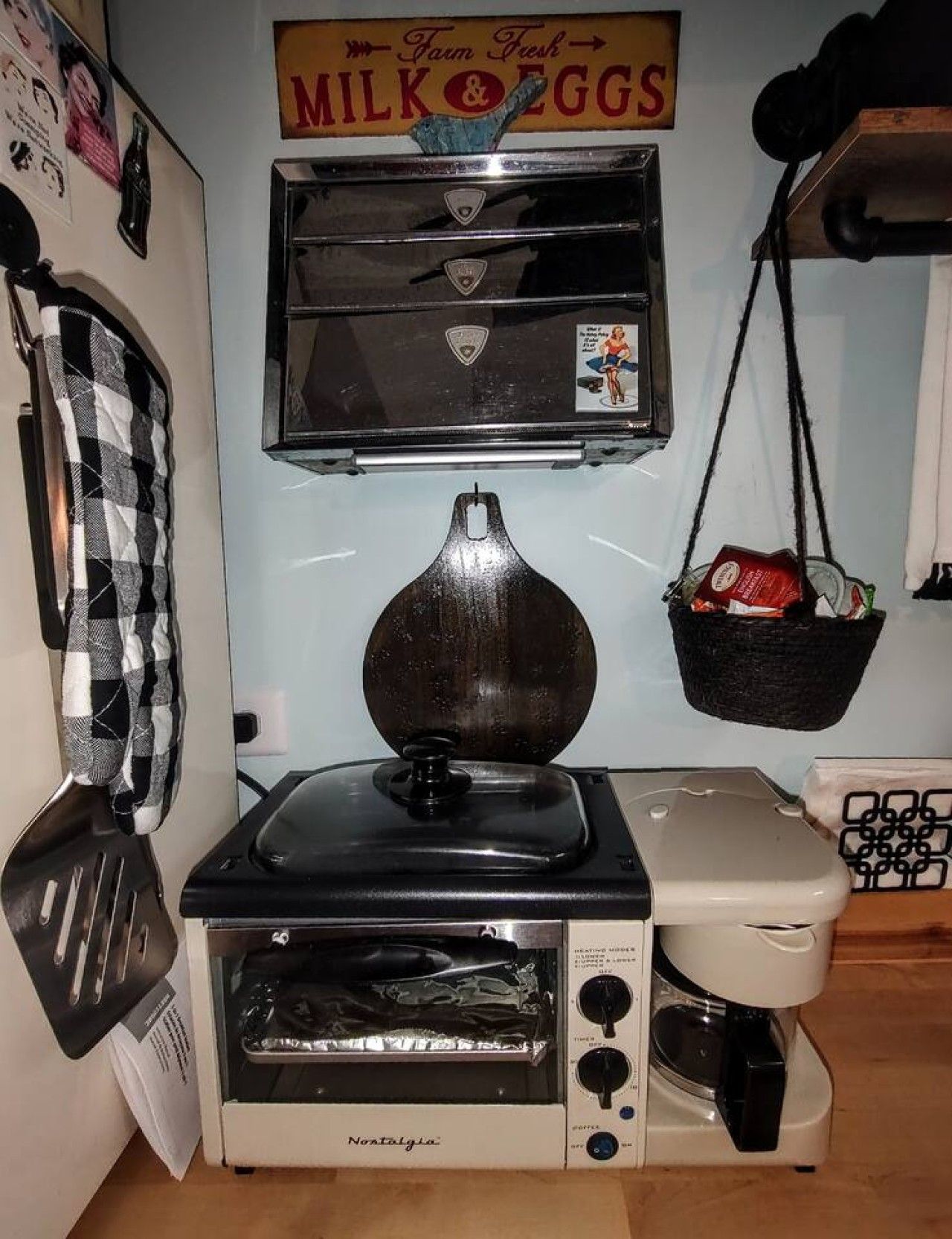 A kitchenette with a vintage toaster, a coffee maker, and a hanging basket with groceries.