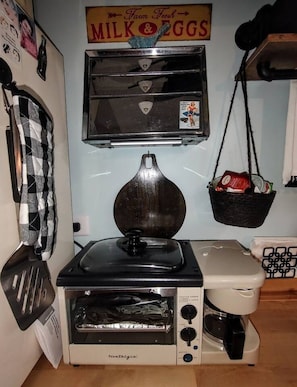 A kitchenette with a vintage toaster, a coffee maker, and a hanging basket with groceries.