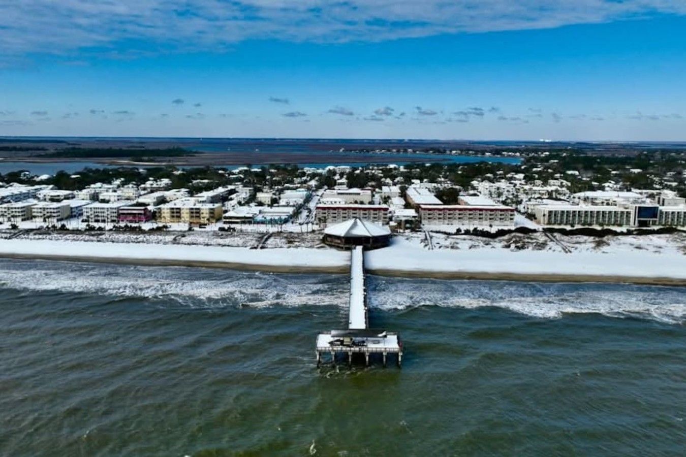 An aerial view of a coastal area with a pier extending into the water, surrounded by buildings and snow-covered ground.