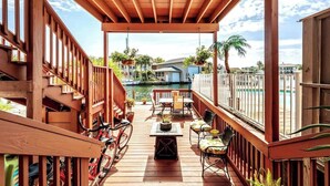 Une terrasse en bois avec un espace salon couvert, un vélo et une vue sur une marina avec des bateaux et des maisons.