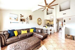 A spacious living room with a brown leather sectional sofa, a ceiling fan, and a kitchen in the background.